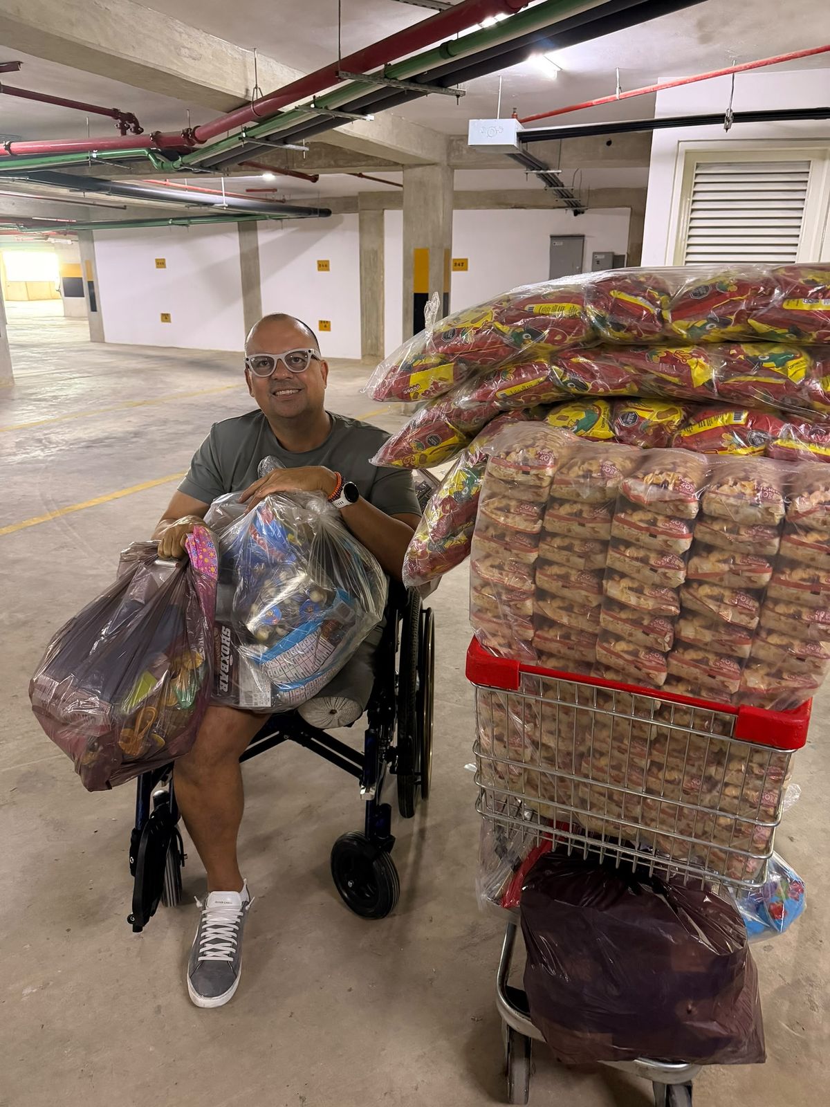 Hombre en silla de ruedas con bolsas de suministros en las piernas, junto a un carrito de compras lleno de paquetes de comida en un estacionamiento techado.