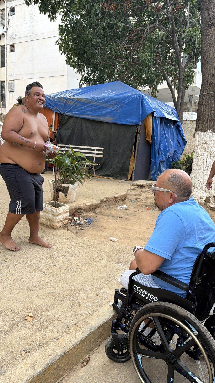 Un hombre en silla de ruedas frente a un hombre de pie, junto a una carpa azul en un terreno de tierra.
