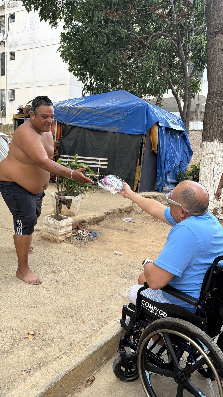 Un hombre en silla de ruedas entrega un paquete a un hombre de pie junto a una carpa azul.