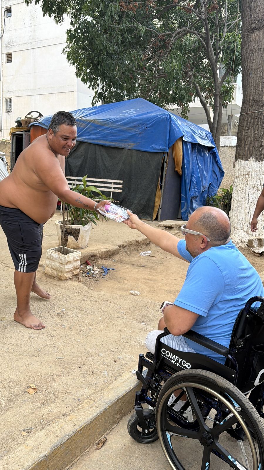 Un hombre en silla de ruedas entrega un paquete a un hombre de pie frente a una carpa azul.