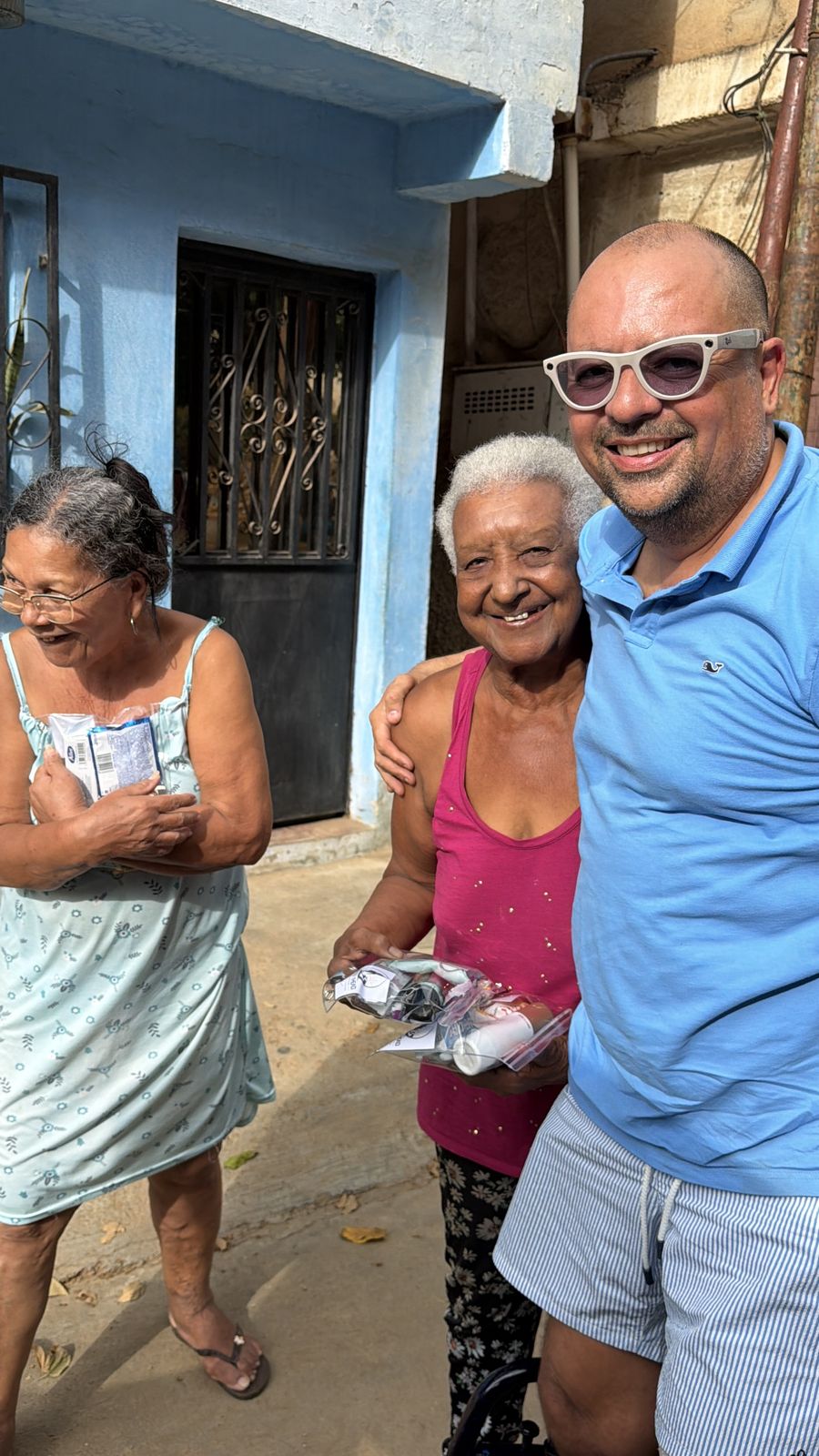 Dos señoras mayores, una sosteniendo una bolsa de ayuda, posan junto a un hombre con lentes de sol frente a una casa.